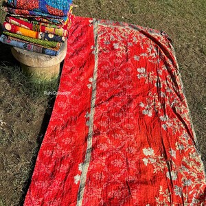 May include: A red kantha quilt with a floral pattern and a white border. The quilt is folded in half and lying on a bed of green grass.