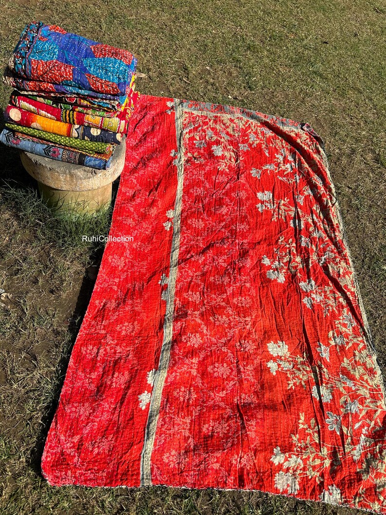 May include: A red kantha quilt with a floral pattern and a white border. The quilt is folded in half and lying on a bed of green grass.
