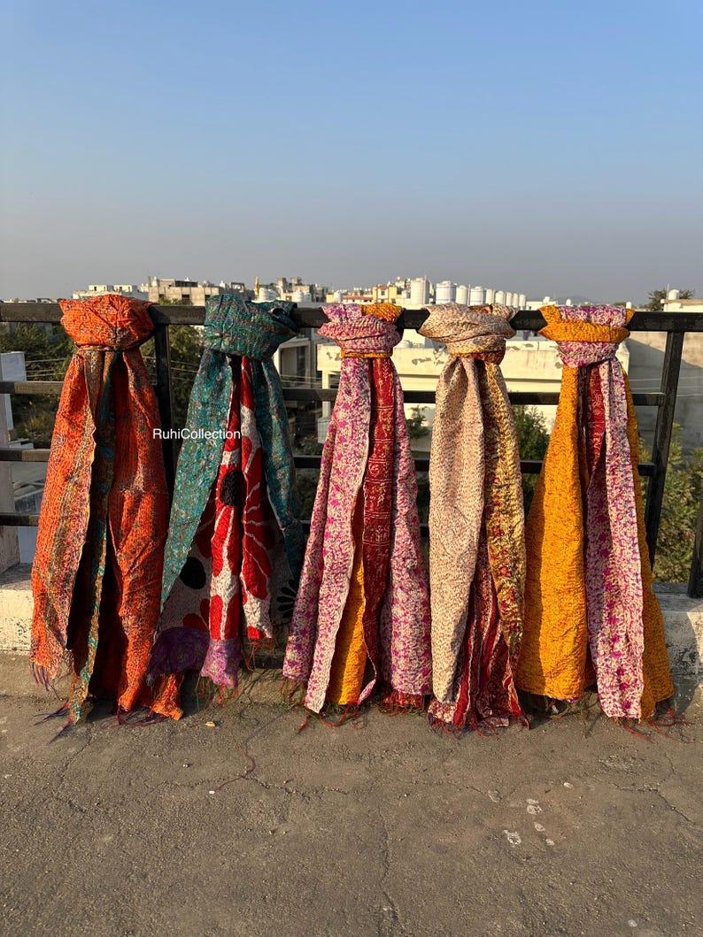 May include: Five colorful kantha quilts are hanging on a railing. The quilts are made of recycled saris and have a variety of patterns and colors, including orange, teal, pink, and yellow.