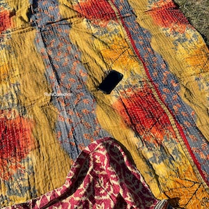 May include: A colorful kantha quilt with a floral pattern in shades of yellow, orange, red, and blue. The quilt is made of recycled saris and has a unique patchwork design. The quilt is folded in half and has a small black phone on top of it.