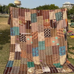 Puede incluir: Un edredón de retazos con una variedad de telas marrones, beige y turquesas. El edredón está hecho de parches rectangulares con patrones florales y geométricos. El edredón se exhibe al aire libre sobre césped.