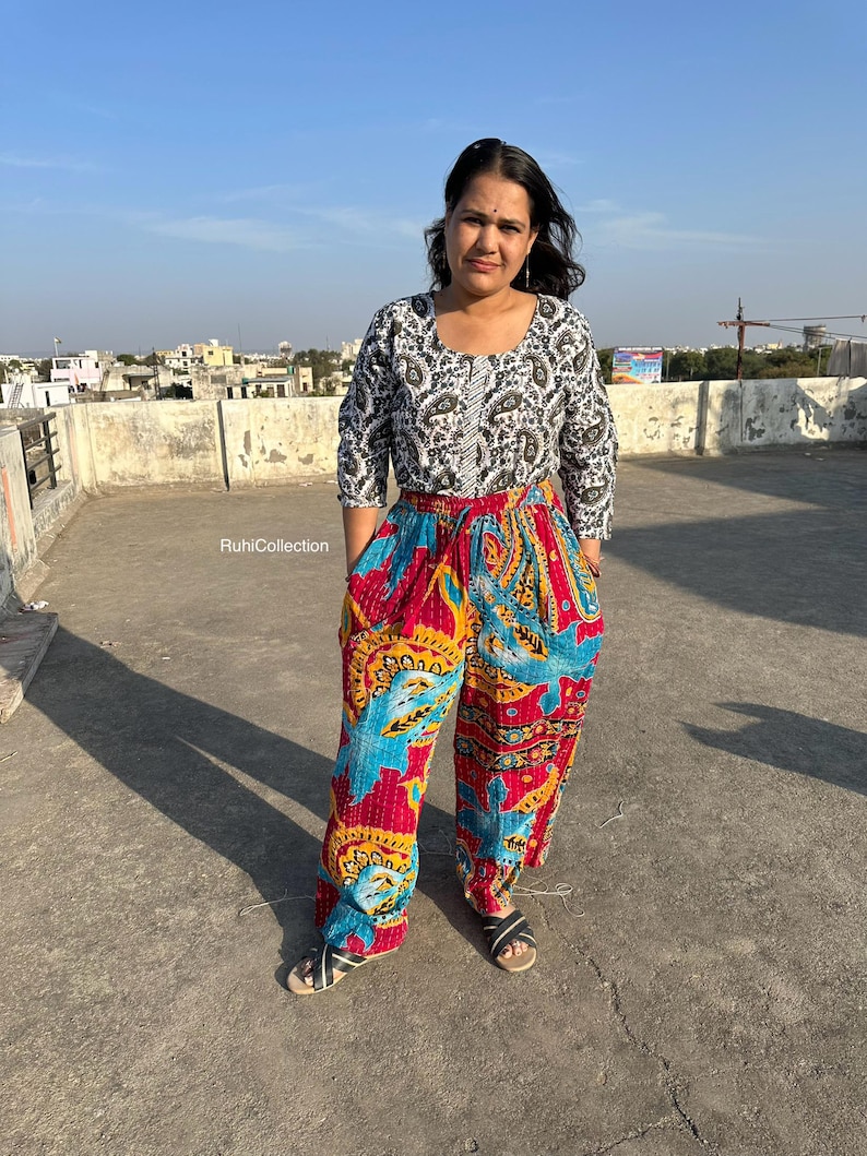 May include: A woman wearing a white and black patterned top with a red, blue, and yellow patterned pair of pants. The pants have a drawstring waist and a wide leg.