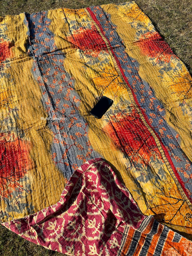 May include: A colorful kantha quilt with a floral pattern in shades of yellow, orange, red, and blue. The quilt is made of recycled saris and has a unique patchwork design. The quilt is folded in half and has a small phone on top of it.