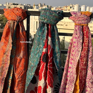 May include: Three colorful kantha scarves hanging on a railing. The scarves are made of recycled saris and feature a variety of floral and geometric patterns. The scarves are in shades of orange, teal, and pink.