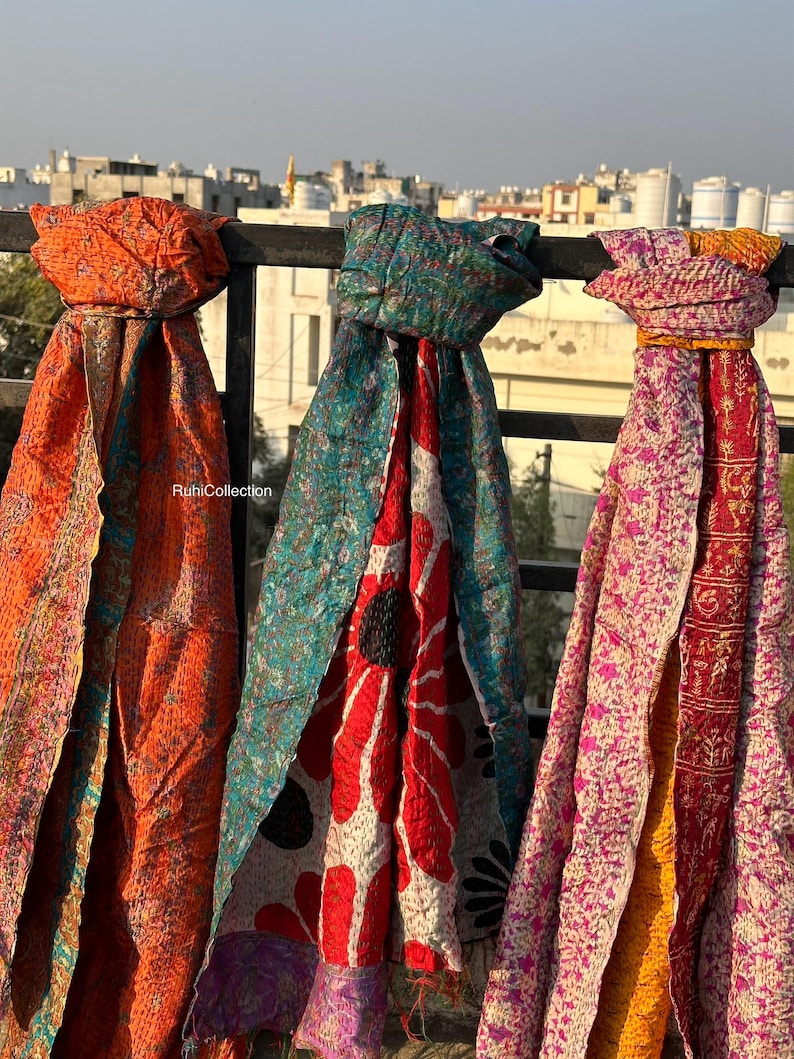 May include: Three colorful kantha scarves hanging on a railing. The scarves are made of recycled saris and feature a variety of floral and geometric patterns. The scarves are in shades of orange, teal, and pink.