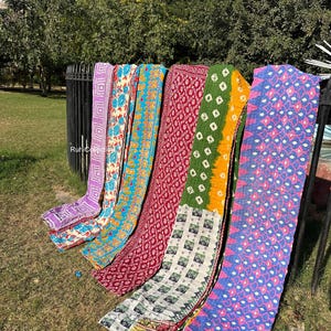May include: Six colorful kantha quilts with various patterns and designs, including floral, geometric, and paisley motifs. The quilts are arranged in a row, hanging from a metal fence. The quilts are made of recycled saris and are a popular choice for home decor and gifts.