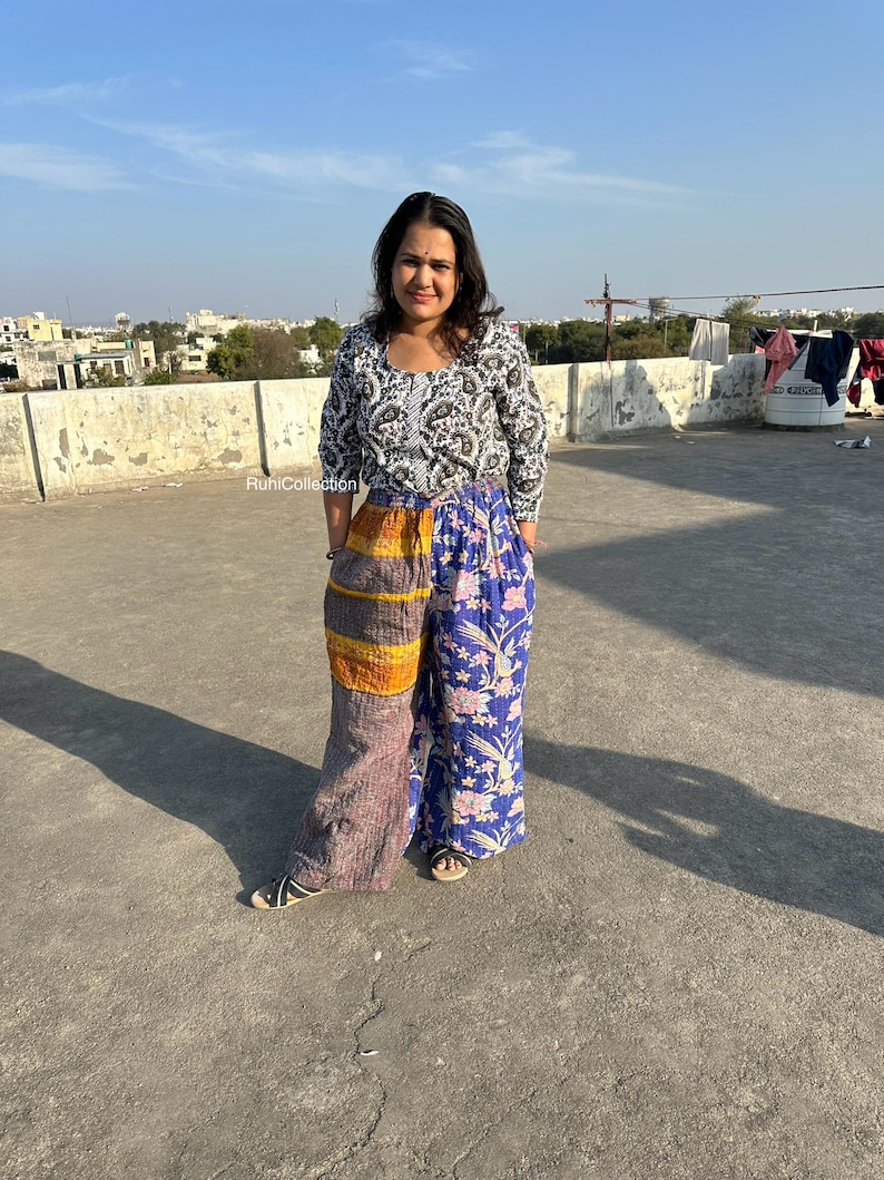 May include: A woman wearing a white and black patterned top with long sleeves and a pair of patchwork pants. The pants are a mix of brown, orange, yellow, and blue fabrics with floral patterns. The woman is standing on a rooftop with a city skyline in the background.