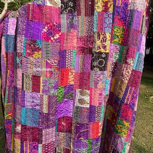 May include: A patchwork quilt in shades of purple, pink, and red. The quilt is made of various fabric squares with different patterns and textures. The stitching is visible, adding to the handmade aesthetic.