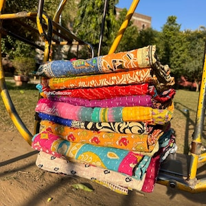 May include: A stack of colorful kantha quilts, made from recycled saris, with intricate patterns and embroidery. The quilts are folded and stacked on a yellow metal swing set.