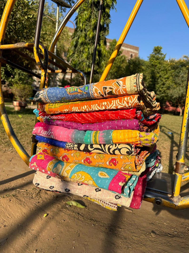 May include: A stack of colorful kantha quilts, made from recycled saris, with intricate patterns and embroidery. The quilts are folded and stacked on a yellow metal swing set.