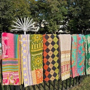 May include: Six colorful kantha quilts hanging on a black metal fence. The quilts are made of patchwork fabric and have a variety of patterns, including floral, geometric, and paisley. The quilts are a vibrant mix of pink, green, yellow, orange, and blue.