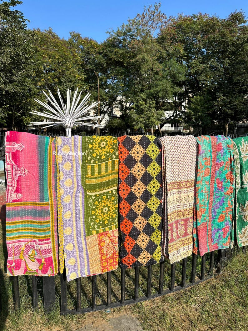 May include: Six colorful kantha quilts hanging on a black metal fence. The quilts are made of patchwork fabric and have a variety of patterns, including floral, geometric, and paisley. The quilts are a vibrant mix of pink, green, yellow, orange, and blue.