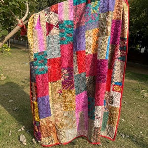 May include: A colorful patchwork quilt with a red border, displayed outdoors. The quilt is made of various fabric squares in shades of pink, red, purple, yellow, and green. The quilt is hanging on a tree branch. The text "RubiCollection" is visible.