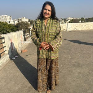 Puede incluir: Una mujer con una chaqueta verde estampada y pantalones de pierna ancha con estampado de leopardo. La chaqueta tiene un diseño floral y los pantalones son marrones y beige. El atuendo se usa al aire libre en un día soleado.