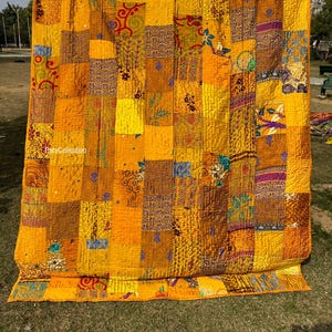 Może przedstawiać: Duża, patchworkowa kołdra w odcieniach żółtego, złotego i brązowego. Kołdra wykonana jest z prostokątnych kawałków materiału o różnych wzorach i fakturach. Kołdra wisi na zewnątrz, z widocznym tekstem "Ruhicollection".