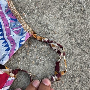 Może przedstawiać: Tekstylny przedmiot typu patchwork z paskiem, prezentujący mieszankę wzor&oacute;w i kolor&oacute;w, w tym niebieski, r&oacute;żowy i brązowy. Pasek wykonany jest z wzorzystej tkaniny. Przedmiot jest trzymany przez dłoń.
