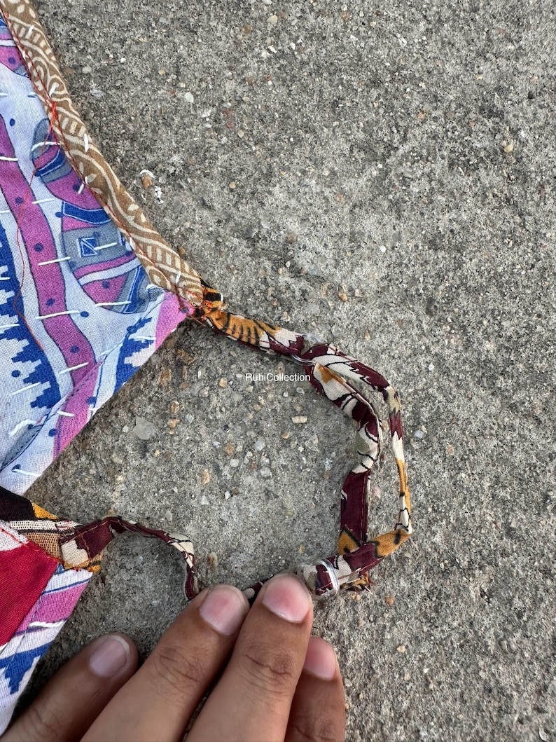 Może przedstawiać: Tekstylny przedmiot typu patchwork z paskiem, prezentujący mieszankę wzor&oacute;w i kolor&oacute;w, w tym niebieski, r&oacute;żowy i brązowy. Pasek wykonany jest z wzorzystej tkaniny. Przedmiot jest trzymany przez dłoń.
