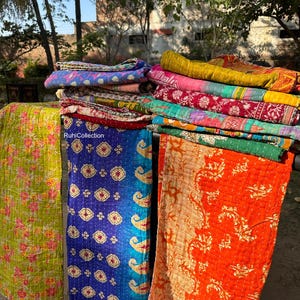 May include: A stack of colorful kantha quilts with floral and paisley patterns. The quilts are made of recycled saris and are hand-stitched. The quilts are hanging on a clothesline in an outdoor setting.