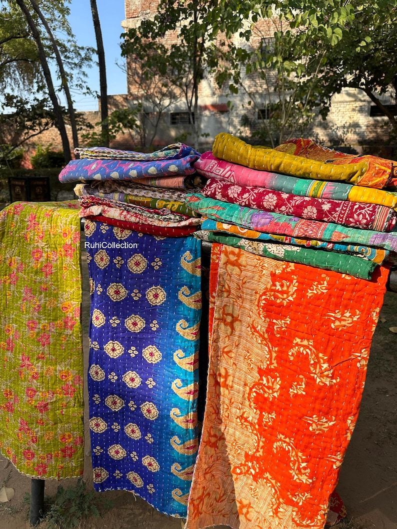 May include: A stack of colorful kantha quilts with floral and paisley patterns. The quilts are made of recycled saris and are hand-stitched. The quilts are hanging on a clothesline in an outdoor setting.