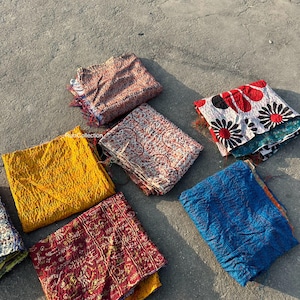 May include: A collection of eight colorful kantha quilts, a type of Indian patchwork textile, arranged on a gray concrete surface. The quilts are made from recycled saris and feature a variety of patterns and colors, including yellow, red, blue, and white.