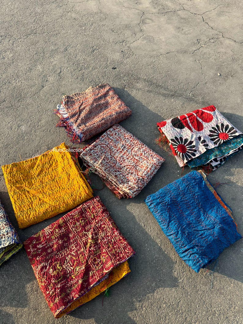 May include: A collection of eight colorful kantha quilts, a type of Indian patchwork textile, arranged on a gray concrete surface. The quilts are made from recycled saris and feature a variety of patterns and colors, including yellow, red, blue, and white.