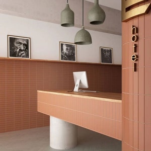 Könnte beinhalten: Ein Hotelrezeptionstresen mit einer Holztheke und einem Computermonitor. Der Tresen ist aus Terrakottafliesen gefertigt und steht vor einer Wand mit Terrakottafliesen. Die Wand hat drei schwarz-weiße gerahmte Fotografien. Die Wand hinter dem Tresen trägt ein goldenes Schild mit der Aufschrift "Hotel".