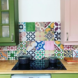 May include: A kitchen with green and pink cabinets, a black electric hob, and a colourful tiled backsplash. Two black pots are on the hob.