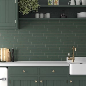 May include: A kitchen with green cabinets and a green tiled backsplash. The backsplash has a subtle gold stripe running through the center of each tile. The countertop is white and there is a white porcelain sink with a brass faucet.
