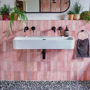 Puede incluir: Un lavabo de porcelana blanca con grifos negros y un tubo de desagüe negro. El lavabo está montado en una pared alicatada con azulejos rectangulares rosas. El suelo está alicatado con pequeñas piedras negras y blancas.