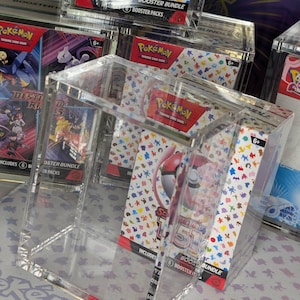 Può includere: Espositori trasparenti in acrilico che contengono pacchetti booster del gioco di carte collezionabili Pokémon. I pacchetti sono etichettati "Scarlet & Violet 151" e "Destiny Rises". Gli espositori sono su una superficie con il logo Pokémon.
