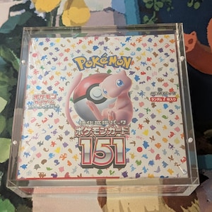 Może przedstawiać: Przezroczysta akrylowa gablota z opakowaniem kart Pokémon. Opakowanie kart zawiera logo Pokémon, różowego Mew i Poké Ball. Tło pokryte jest kolorowymi postaciami Pokémon i numerem 151.