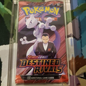 Könnte beinhalten: Versiegeltes Pokémon Scarlet & Violet Destined Rivals Sammelkartenpack. Das Pack zeigt ein lila Mewtwo und einen Mann im schwarzen Anzug. Das Pack ist in einem transparenten Kunststoffgehäuse eingeschlossen.
