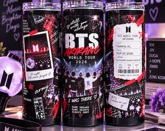 Op de afbeelding: Drie zwarte cilindrische bekers met deksels en rietjes, voorzien van BTS World Tour Arirang 2026 graphics, concertfoto's, een nep-ticket voor Tampa, FL, en teksten zoals Still with you always, I was there en Same dreams same hearts.