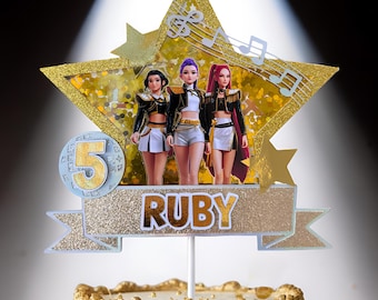 May include: A cake topper featuring three dolls in black and white outfits, standing in front of a gold star with musical notes. The topper has the word "RUBY" and the number "5" on it. The cake itself is gold with ornate details.