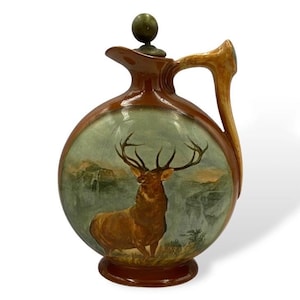 May include: A round ceramic decanter with a brown glazed finish, featuring a painted scene of a stag in a landscape on the front. The vessel has a curved handle, a short spout, and a spherical brass-toned stopper on top of the lid.