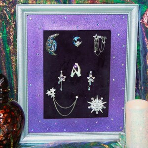 May include: A silver frame with a purple glitter background displays a collection of celestial-themed jewellery. The jewellery includes stars, moons, and a pair of hands holding a crystal.