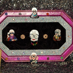 May include: An octagonal pink frame with a silver glitter border and black velvet interior.  Four images of women wearing glasses and colorful clothing are displayed in the frame.  Two gold-colored decorative accents are on either side of the center image.