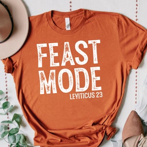Może przedstawiać: Pomarańczowy t-shirt z napisem "FEAST MODE" w kolorze białym z przetartym fontem. Pod tekstem znajduje się "LEVITICUS 23" mniejszym fontem.