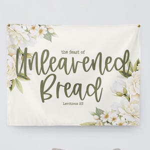 May include: A cream-colored wall hanging with the text "the feast of Unleavened Bread" in a script font, with "Leviticus 23" below. White floral arrangements with green leaves adorn the corners.