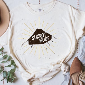 Może przedstawiać: Biała koszulka z brązowym nadrukiem namiotu sukkot i tekstem "Sukkot Mode". Nadruk jest otoczony żółtymi promieniami słońca.