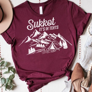 Może przedstawiać: Bordowy t-shirt z białym nadrukiem graficznym przedstawiającym góry i namiot. Tekst "Sukkot It's in Tents" jest napisany białym pismem ozdobnym.