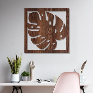 May include: A square wooden wall art piece featuring a cut-out design of a monstera leaf. The leaf is brown with visible wood grain, set against a white background. The artwork is framed in a dark brown border, creating a striking contrast.