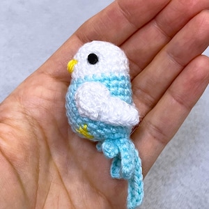 Op de afbeelding: Een kleine, handgemaakte gehaakte vogel in wit, lichtblauw en geel. De vogel heeft een zwart oog en een gele snavel. De vleugels zijn wit en het lichaam is lichtblauw met witte accenten. De staart is lichtblauw. Een charmant, handgemaakt item.
