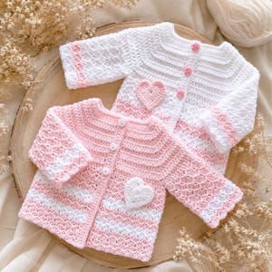 May include: Two handmade crochet baby cardigans. One is white with pink accents and a heart applique, the other is pink with white stripes and a heart. Both have button closures and are displayed on a wooden surface.