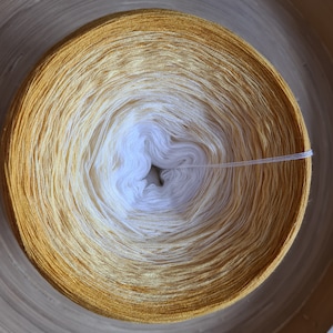 May include: A round skein of yarn with a gradient color scheme, transitioning from a deep golden yellow to a creamy white center. The yarn is tightly wound, showcasing the color variations. The yarn is for knitting and crocheting projects.