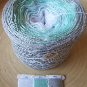 Op de afbeelding: Een grote bol garen met een kleurovergang, waaronder grijs, mintgroen en wit. Er is ook een kleine plastic kaart met drie draadstalen in bijpassende kleuren. Het garen is waarschijnlijk voor brei- of haakprojecten.