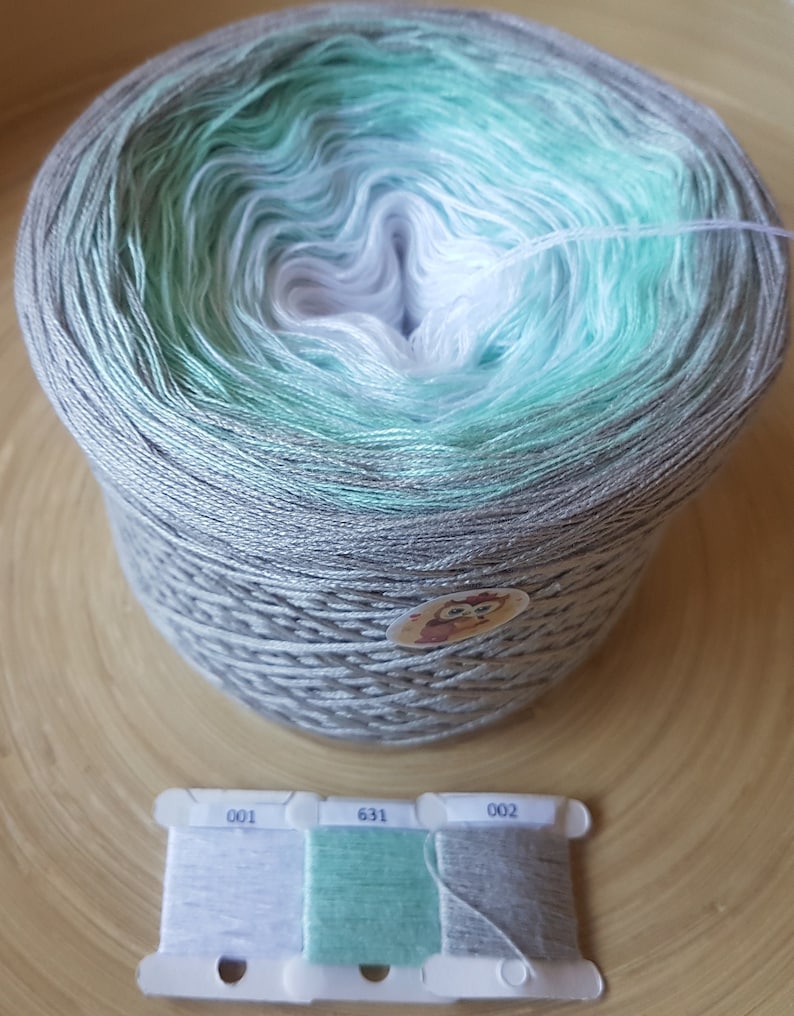 Op de afbeelding: Een grote bol garen met een kleurovergang, waaronder grijs, mintgroen en wit. Er is ook een kleine plastic kaart met drie draadstalen in bijpassende kleuren. Het garen is waarschijnlijk voor brei- of haakprojecten.