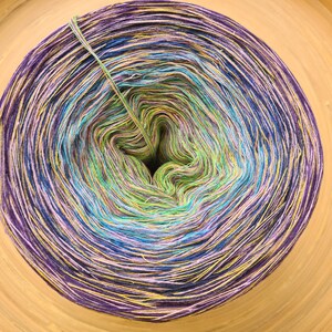 May include: A tightly wound ball of yarn in shades of purple, blue, green, and yellow. The yarn is arranged in concentric circles, with a hole in the center. A single strand of green yarn extends from the ball.