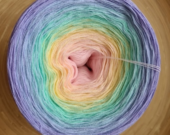 May include: A circular skein of yarn in a gradient of pastel colors, including lavender, light blue, green, yellow, and pink. The yarn is tightly wound, with strands extending from the center. The yarn is likely for knitting or crochet projects.