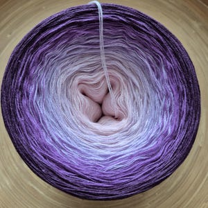 Peut inclure: Pelote de laine aux couleurs dégradées, passant du violet foncé au rose clair. Le fil est bien enroulé, avec un brin qui s'étend du centre. Idéal pour les projets de tricot ou de crochet.